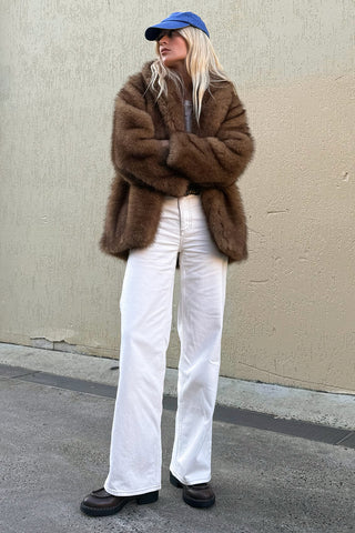 marina-wearing-camille-coat-golden-brown-faux-fur-arms-crossed-styled-with-white-wide-leg-jeans-and-blue-cap