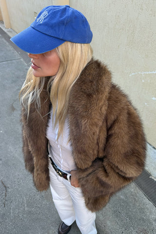 side-view-of-marina-in-golden-brown-faux-fur-coat-with-wide-collar-styled-with-white-jeans-and-blue-cap