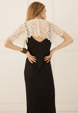 a-woman-shown-from-the-back-wearing-a-black-maxi-dress-with-a-sheer-white-lace-cape-style-shoulder-overlay-standing-against-a-white-paneled-wall