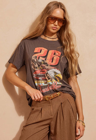 a-woman-wearing-a-faded-brown-graphic-t-shirt-with-a-large-26-and-eagle-print-styled-with-brown-pants-and-sunglasses-against-a-beige-wall