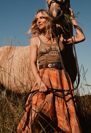A woman in a rustic western-style outfit stands in a grassy field with a horse beside her. She wears a brown tank top with western text graphics and a long rust-orange plaid skirt
