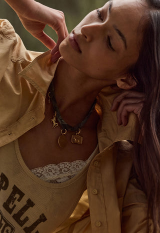 a-close-up-lifestyle-shot-of-a-woman-wearing-a-layered-necklace-set-the-set-includes-a-dark-green-braided-cord-necklace-with-a-small-gold-crescent-moon-and-a-gold-square-charm-layered-over-a-gold-chain-with-a-gold-n-initial-pendant