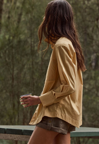 a-woman-shown-from-the-back-wearing-a-loose-long-sleeve-mustard-yellow-button-up-shirt-and-brown-shorts-holding-a-coffee-cup-in-an-outdoor-setting-with-a-blurred-forest-background