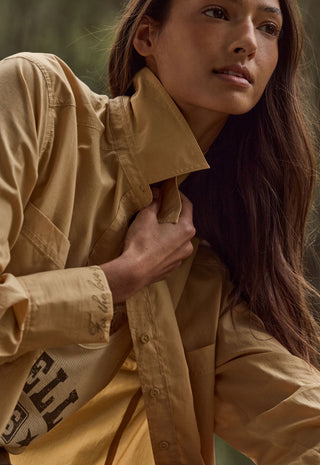 a-close-up-lifestyle-shot-of-a-model-wearing-a-tan-utility-shirt-jacket-with-a-high-collar-and-a-matching-tan-top-underneath-the-jacket-features-a-chest-pocket-and-a-sleeve-with-a-black-graphic-print