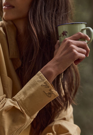 a-close-up-lifestyle-shot-of-a-woman-holding-a-light-green-enamel-mug-with-a-dark-rim-and-palm-tree-print-wearing-a-tan-button-up-shirt-with-embroidered-script-on-the-cuff