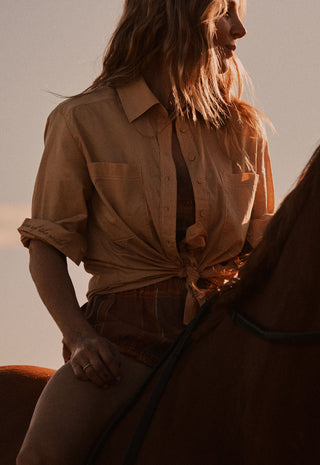 a-woman-wearing-a-tan-button-up-shirt-tied-at-the-waist-sits-on-a-horse-in-warm-golden-hour-light
