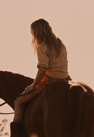 a-woman-with-long-light-brown-hair-sits-on-a-horse-shown-from-behind-in-warm-backlit-outdoor-lighting-she-wears-a-beige-top-and-orange-plaid-shorts-against-a-solid-pale-peach-sky