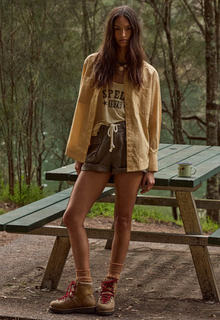 a-woman-stands-outdoors-at-a-wooden-picnic-table-in-a-wooded-setting-wearing-a-tan-overshirt-a-graphic-t-shirt-brown-shorts-and-tan-lace-up-boots