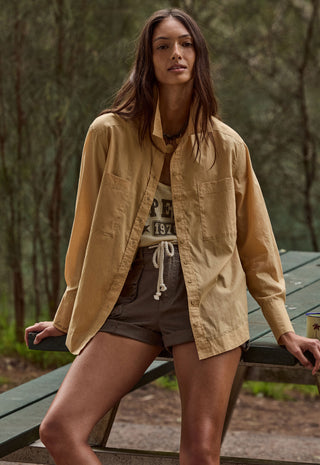 a-woman-wearing-a-tan-long-sleeve-button-up-shirt-over-a-graphic-tee-and-brown-shorts-leaning-on-a-green-picnic-table-in-a-wooded-outdoor-setting