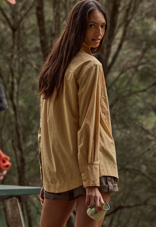 a-woman-wearing-an-oversized-tan-long-sleeve-shirt-and-dark-shorts-photographed-outdoors-against-a-blurred-forest-background