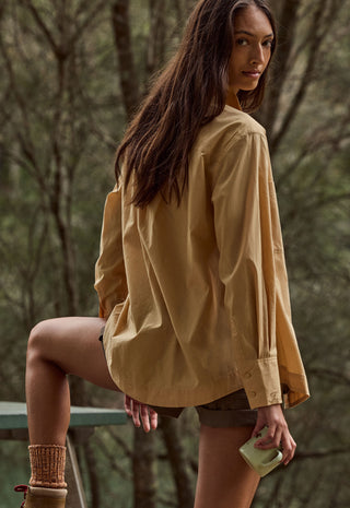 a-woman-wearing-an-oversized-tan-button-up-shirt-and-brown-shorts-posing-outdoors-in-a-wooded-setting