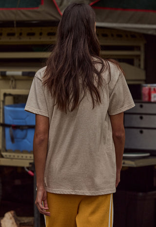 A person viewed from behind wearing an oversized light beige short-sleeve t-shirt and mustard yellow pants, standing in front of a rustic outdoor market stall.