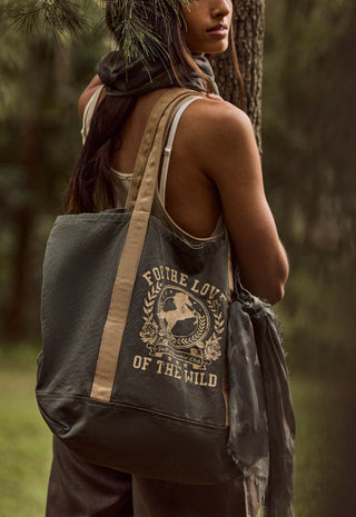 a-woman-outdoors-by-a-tree-wearing-a-dark-olive-canvas-tote-bag-with-beige-straps-and-a-distressed-cream-graphic-that-reads-for-the-love-of-the-wild