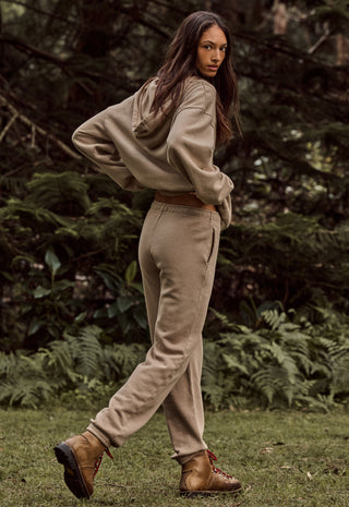 a-woman-wearing-a-beige-cropped-hoodie-and-matching-jogger-pants-with-brown-boots-standing-outdoors-in-a-grassy-area-with-a-forest-background
