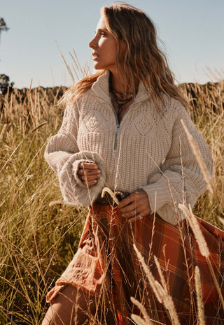 A woman standing in a field of dry golden grass, wearing a cream cable-knit half-zip sweater and a rust-orange plaid skirt.