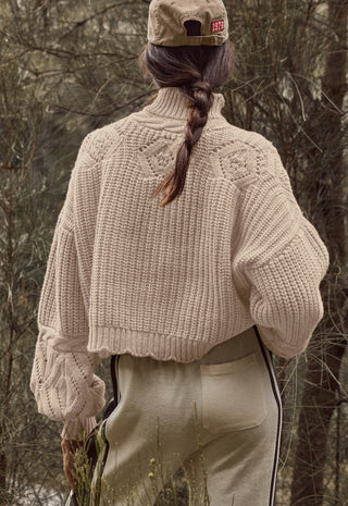A person shown from behind wearing a cream-colored chunky knit cropped sweater, beige pants, and a beige cap, standing outdoors in a wooded setting.