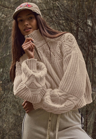 A woman wearing a chunky cable-knit turtleneck sweater in a warm cream/beige color, styled with a matching beige baseball cap and beige sweatpants, set against a blurred forest background.
