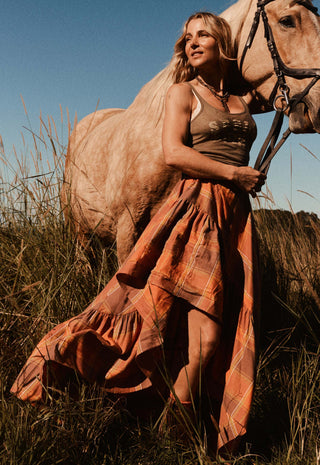 A woman in a rust-orange plaid maxi dress stands outdoors in tall grass beside a light-colored horse