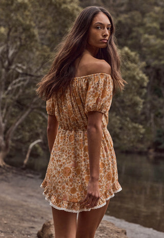 A woman wearing an off-the-shoulder mini dress with a rust-orange and beige floral pattern, standing outdoors near a body of water with trees in the background.