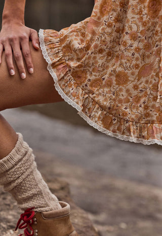 Close-up of a person wearing a tan and brown floral skirt with a white lace hem, paired with beige knit leg warmers and brown boots with red laces.