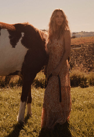 A woman in a sleeveless, patterned maxi dress stands in a grassy field next to a brown and white horse, with a warm golden sunset background.