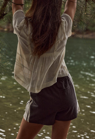 a-woman-viewed-from-behind-wearing-a-loose-off-white-short-sleeve-top-and-dark-charcoal-shorts-with-white-side-stripes-standing-outdoors-near-water-and-hanging-tree-branches