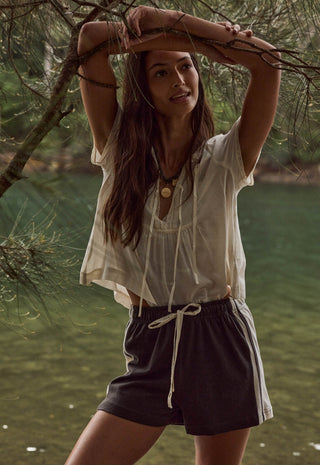 a-woman-outdoors-by-a-body-of-water-under-tree-branches-wearing-a-loose-off-white-short-sleeve-blouse-and-dark-charcoal-drawstring-shorts-with-white-side-stripes