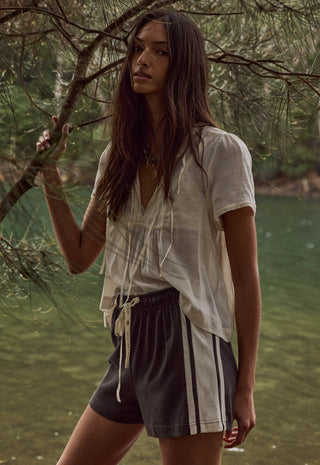 a-woman-standing-outdoors-near-water-and-tree-branches-wearing-a-loose-white-short-sleeve-top-and-dark-gray-drawstring-shorts-with-white-side-stripes