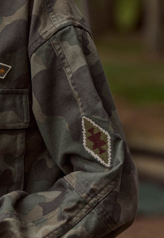 close-up-of-a-camouflage-jacket-sleeve-with-a-diamond-shaped-embroidered-patch-featuring-a-red-star-and-white-border-worn-outdoors-with-a-blurred-background