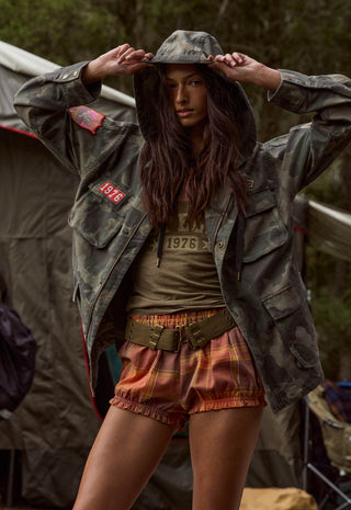 a-woman-wearing-a-vintage-style-military-camouflage-jacket-over-an-olive-green-graphic-t-shirt-paired-with-orange-plaid-shorts-and-a-wide-belt-posing-in-front-of-a-tent-outdoors
