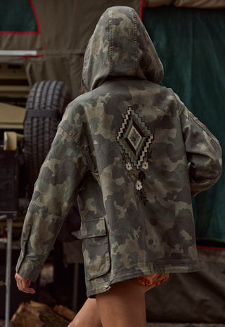 a-person-viewed-from-behind-wearing-a-hooded-camouflage-jacket-with-a-large-geometric-diamond-pattern-and-small-white-flower-details-on-the-back-set-against-a-dark-outdoor-backdrop