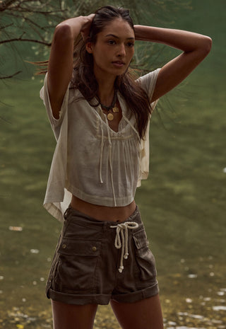 a-woman-standing-outdoors-near-water-wearing-a-loose-short-sleeve-cream-top-and-dark-brown-cargo-shorts-with-a-white-drawstring