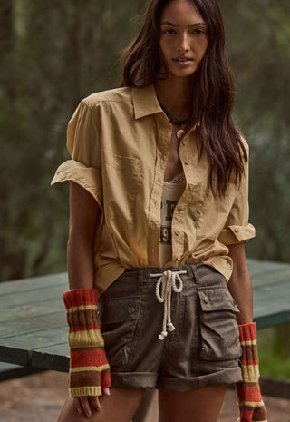 a-woman-wearing-a-tan-button-up-shirt-brown-cargo-shorts-and-striped-knit-fingerless-gloves-standing-outdoors-in-front-of-a-blurred-forest-background
